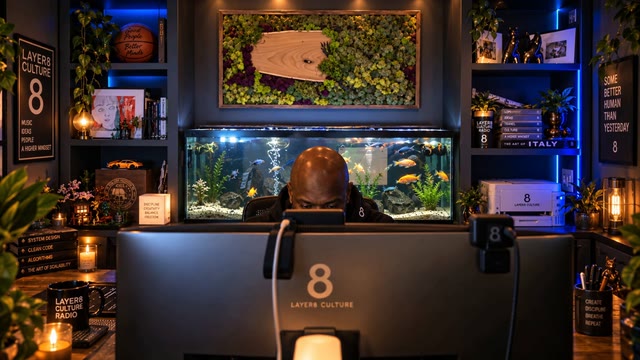 Layer8Culture Radio studio with a builder at a workstation, an illuminated aquarium, and warm blue-and-gold lighting